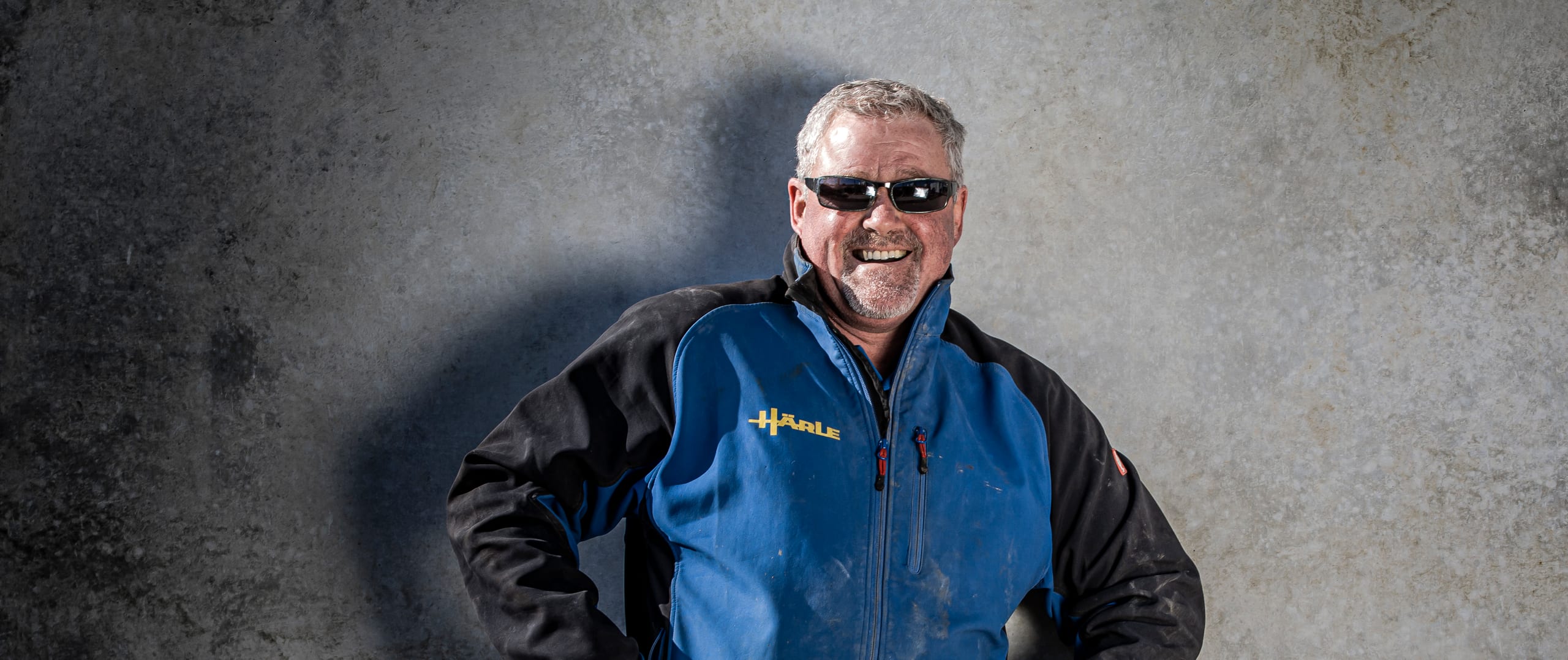 Lachender Mitarbeiter von Härle Bau mit Sonnenbrille in Härle-Arbeitsjacke vor einer Betonwand
